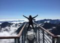 person standing on hand rails with arms wide open facing the mountains and clouds